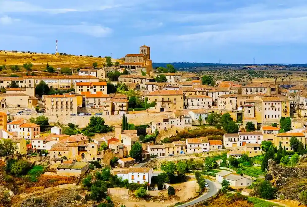 Uma vista panorâmica aérea da vila medieval de Sepúlveda, em Castela e Leão, Espanha, aninhada numa colina de pedra dourada com o desfiladeiro das Hoces del Duratón e campos rurais que se estendem ao fundo sob um céu nublado.