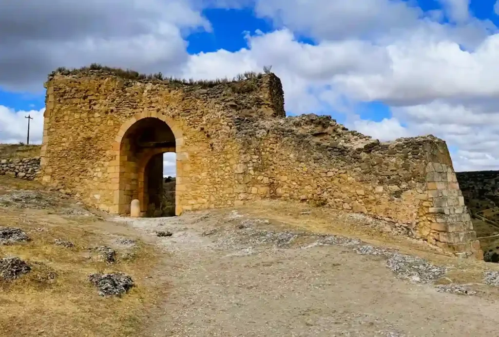 As ruínas de pedra da Puerta de la Fuerza em Sepúlveda, uma das sete portas medievais originais, com um arco rústico e paredes em alvenaria de pedra, situadas num terreno árido sob um céu azul com nuvens.