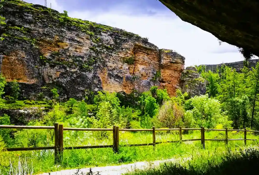 Vista de um trilho de terra no Parque Natural das Hoces del Río Duratón em Sepúlveda, delimitado por uma cerca de madeira e vegetação verdejante, com o imponente desfiladeiro de paredes de pedra vertical e colorida ao fundo sob um céu azul com nuvens.