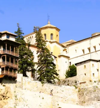 Fachada em perspetiva das Casas Colgadas de Cuenca com as suas icónicas varandas de madeira medievais suspensas sobre a rocha calcária sob um céu azul.