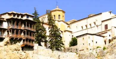 Fachada em perspetiva das Casas Colgadas de Cuenca com as suas icónicas varandas de madeira medievais suspensas sobre a rocha calcária sob um céu azul.