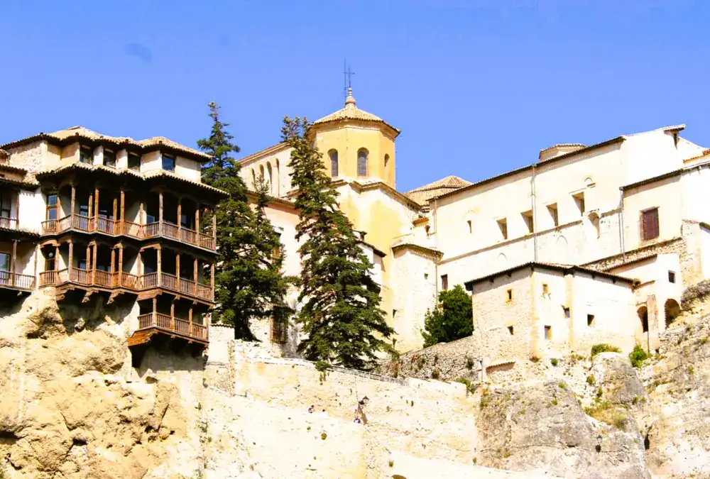 Fachada em perspetiva das Casas Colgadas de Cuenca com as suas icónicas varandas de madeira medievais suspensas sobre a rocha calcária sob um céu azul.