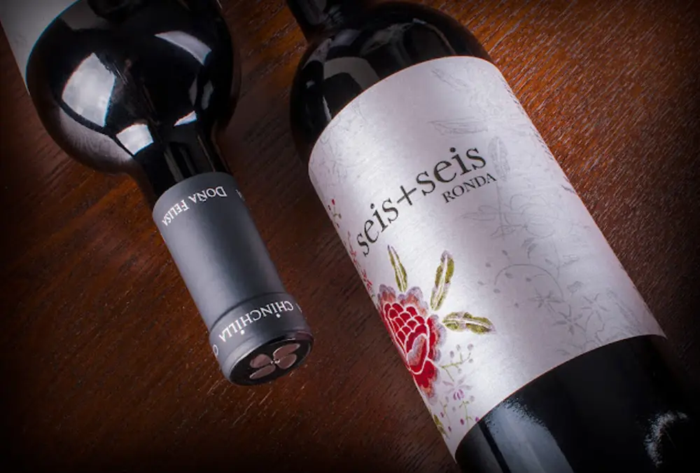 Duas garrafas de vinho tinto Seis mais Seis da Bodega Doña Felisa de Ronda deitadas sobre mesa de madeira escura.
