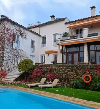 Piscina azul com espreguiçadeiras junto a uma parede de pedra e fachada com plantas.