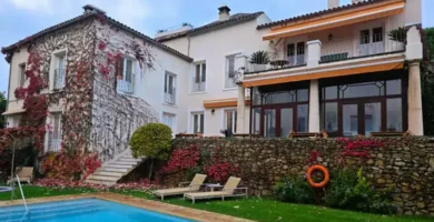 Piscina azul com espreguiçadeiras junto a uma parede de pedra e fachada com plantas.