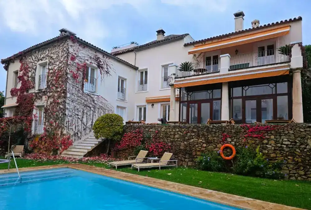 Piscina azul com espreguiçadeiras junto a uma parede de pedra e fachada com plantas.