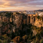 Vista panorâmica do desfiladeiro de Ronda e da Puente Nuevo ao pôr-do-sol, com o casario branco no topo das arribas rochosas.